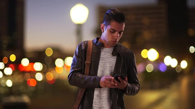 Millennial Hispanic man standing on city bridge at night looking at smartphone. Latino guy on urban street in the evening using cell phone