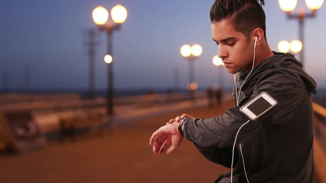 Millennial Hispanic Jogger checking fitness tracker while resting on boardwalk. Latino runner jogging on pier listening to music with smartphone and earbuds