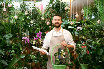 Handsome florist holding basket with flower and notebook in greenhouse