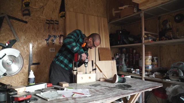 profession, people, carpentry, woodwork and people concept - carpenter with electric drill drilling wood plank at workshop