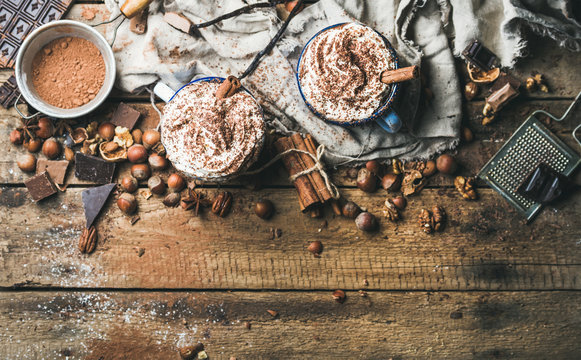 Hot chocolate with whipped cream and cinnamon sticks served with anise, different nuts and cocoa powder on rustic wooden background, top view, selective focus, copy space, horizontal composition