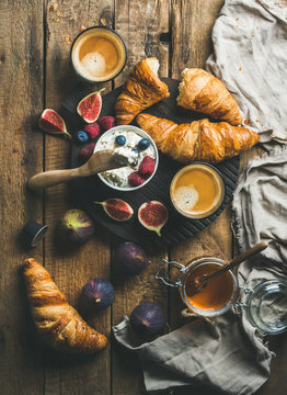 Breakfast with croissants, homemade ricotta cheese, figs, fresh berries, honey and glasses of espresso coffee on dark serving board over rustic wooden background, top view, vertical composition