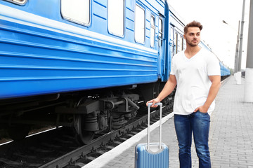 Young man in blank t-shirt with suitcase standing on railway platform near train