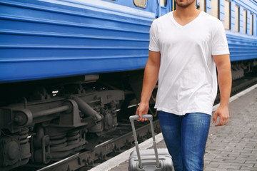 Young man in blank t-shirt walking along railway platform with suitcase, closeup