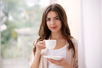 Beautiful woman with cup of aromatic coffee standing near window