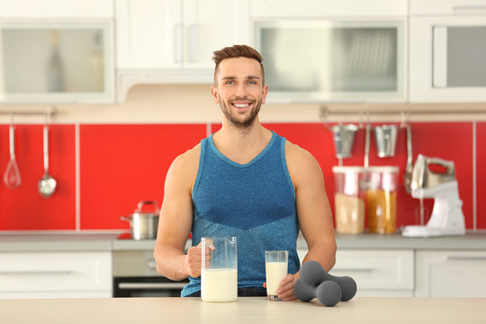 Young sporty man with jug, glass of fresh milk and dumbbells at kitchen
