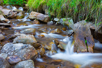 Naklejka na ściany i meble Mountain stream(creek) in the stones and green grass banks in mountain forest. Crystal clear water - rare condition of modern enviroment. Long exposure.