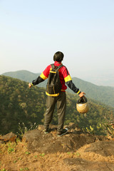 Young man with backpack and helmet standing with raised hands on top of a mountain and enjoying valley view