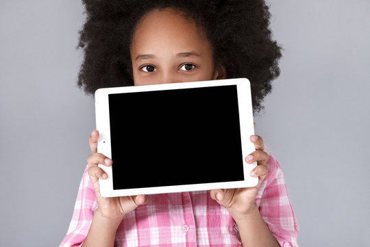 Pretty little girl holding tablet and looking at camera while standing against grey background. Copy space