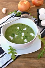 Green soup from arugula and garlic with goat cheese in white bowl with spoon on wooden background