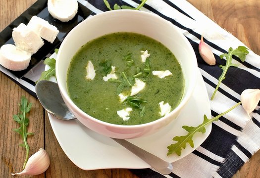 Green soup from arugula and garlic with goat cheese in white bowl with spoon on wooden background