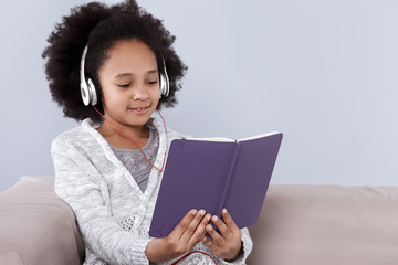 Pretty little girl in headphones holding notebook and smiling while sitting on sofa
