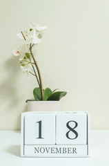 Closeup white wooden calendar with black 18 november word with white orchid flower on white wood desk and cream color wallpaper in room textured background , selective focus at the calendar