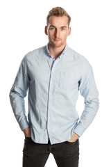 A handsome man in his 20s standing against a white background, smiling, wearing a blue shirt and grey jeans.
