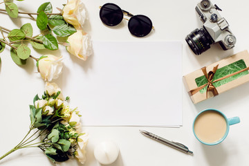 White desk with coffee cup, sunglasses, roses and film camera. Empty sheet in the middle. Top view, flat lay, copyspace.