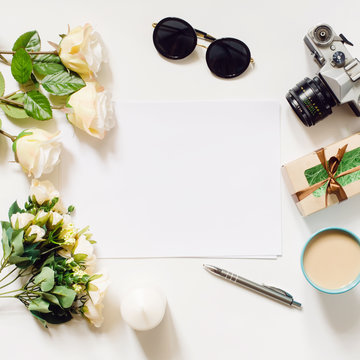 White desk with coffee cup, sunglasses, roses and film camera. Empty sheet in the middle. Top view, flat lay, copyspace.