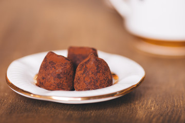 Three candies of truffle on the white plate that stand on a wooden table. On a background there is a cup. Selective focus on a forward part of candies. 