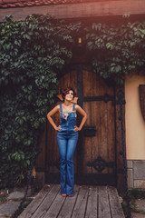Portrait of a beautiful woman wearing denim jumpsuit standing against an ivy framed ancient wooden door of a castle. Magical toned