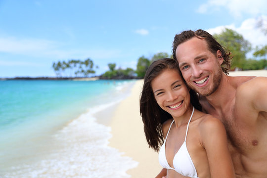 Happy couple selfie on exotic beach vacation. Beautiful interracial young adults smiling taking a self-portrait photo with mobile phone on tropical travel destination with turquoise ocean background.