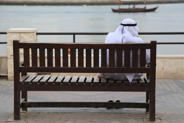 Arab man in dishdasha sitting on a wooden bench at Creek, Dubai - obrazy, fototapety, plakaty