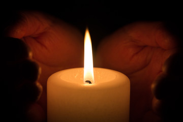 Hände umschließen weiße brennende Kerze im Dunkeln