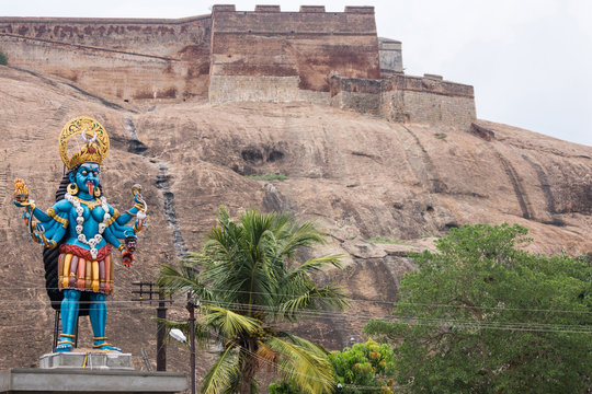 Dindigul, India - October 23, 2013: Large mainly blue colorful bloody cruel goddess Kali statue at the foot of the Rock Fort. Ramparts are backdrop. Green foliage. Beige and browns background.