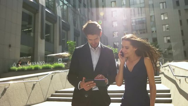 Two young colleagues walking on stairs, woman talking on phone, man using tablet outdoors.