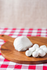 Water buffalo mozzarella mozzarella di buffala cheese in ball and bocconcini balls on wooden cheese chopping board on red and white checkered tableclothbrown burlap background copy space above