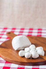 Water buffalo mozzarella mozzarella di buffala cheese in ball and bocconcini balls on wooden cheese chopping board on red and white checkered tableclothbrown burlap background copy space above