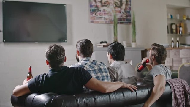 Rear view of male friends sitting on sofa drinking beer and watching game on TV
