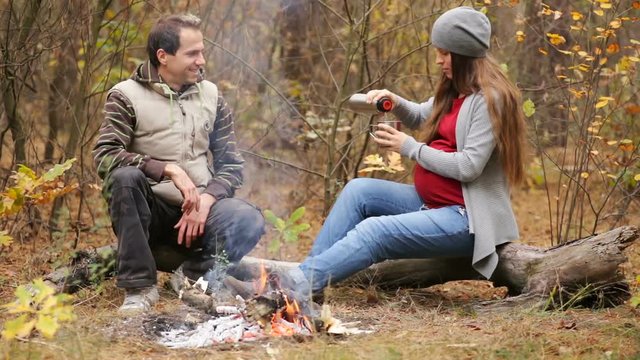 Beautiful  pregnant woman with her husband resting in the forest.  They sit near the fire, a woman pours tea to her husband. The concept of family happiness
