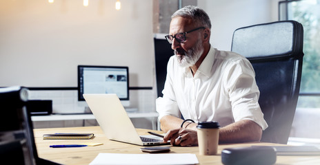 Adult professional businessman wearing a classic glasses and working at the wood table in modern coworking studio.Stylish bearded middle age man using laptop on workplace. Horizontal wide,blurred.