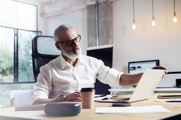 Middle age top manager wearing a classic glasses and working at the wood table in modern interior design office.Stylish bearded businessman using laptop on workplace. Horizontal,blurred.