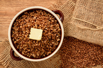 Boiled buckwheat porridge in ceramic pot on wooden background.