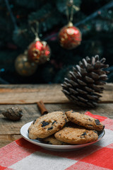 Chocolate chip cookies on rustic background. Nearby lie cinnamon sticks and cones. Behind is tree with toys and garlands. New Year holiday.