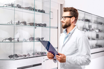 Handsome ophthalmologist with recipe in folder choosing a pair of eyeglasses. Optometrist checking the showcase with glasses in a the store or hospital.