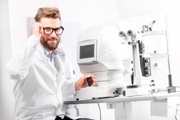Portrait of handsome eye doctor sitting with ophthalmologic device in the cabinet - obrazy, fototapety, plakaty