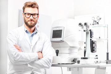 Portrait of handsome eye doctor sitting with ophthalmologic device in the cabinet - obrazy, fototapety, plakaty