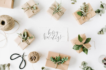 calligraphy words happy holidays and beauty arrangement frame of craft boxes and green branches on white background. flat lay, top view