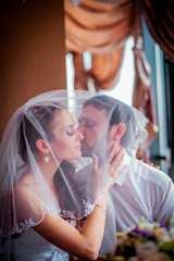 Fototapeta premium Bride kisses on the cheek the groom happy couple holding a bouquet in their hands in the dark interior near the window. Close-up