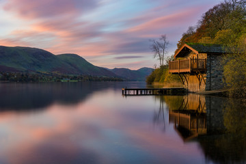 Fototapeta na wymiar Pink and purple vibrant sunrise with dawn light hitting boathouse on calm lake with reflections in the Lake District.