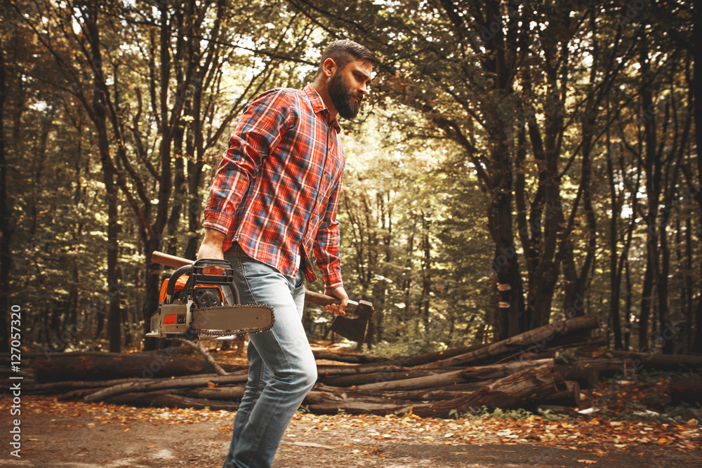 Wall mural lumberjack worker walking in the forest with axe and chainsaw.