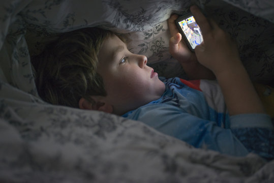 Young boy playing with cell phone under duvet