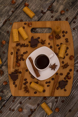 Small white cup of coffee, cinnamon sticks, cocoa beans, star anise, hazelnuts, chocolate, and cookies on wooden background