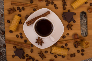 Small white cup of coffee, cinnamon sticks, cocoa beans, star anise, hazelnuts, chocolate, and cookies on wooden background