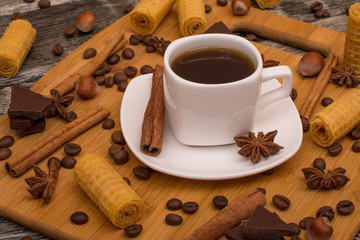 Small white cup of coffee, cinnamon sticks, cocoa beans, star anise, hazelnuts, chocolate, and cookies on wooden background