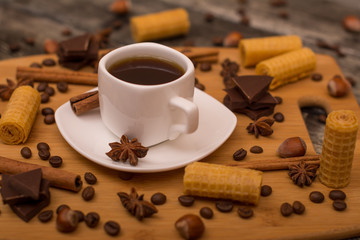 Small white cup of coffee, cinnamon sticks, cocoa beans, star anise, hazelnuts, chocolate, and cookies on wooden background