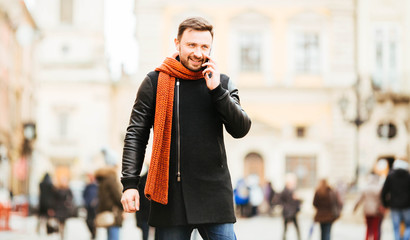 handsome and young man in a black coat speaks by phone outdoors
