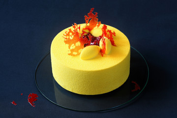 Contemporary Yellow Velvet Mousse Cake decorated with red caramel and wild strawberries, on dark blue background.