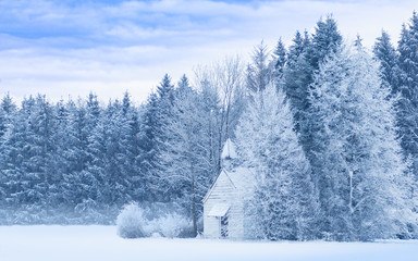Idyllic serene winter landscape with snowy frosty forest and snowbound small woody chapel on...